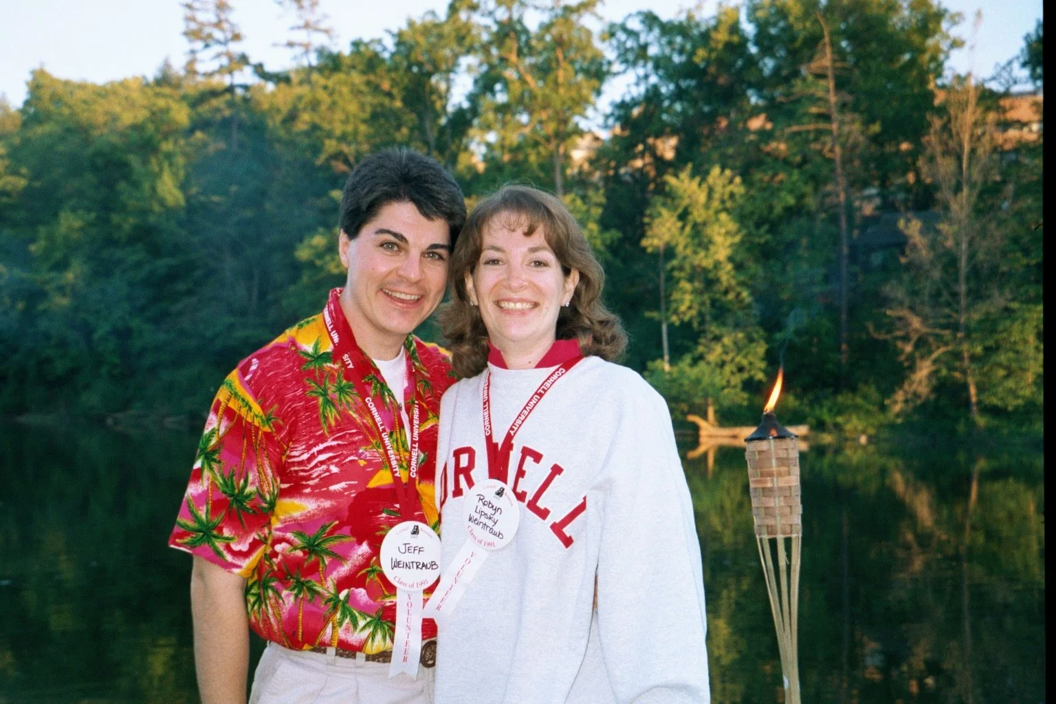 Two people smiling outdoors near a body of water with trees in the background. They are wearing volunteer badges around their necks, and a lit tiki torch is nearby.