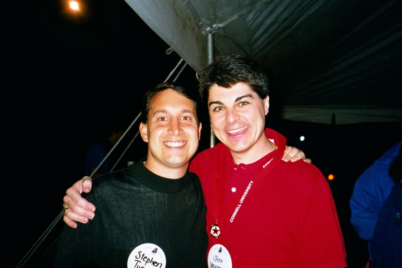 Two men smiling at a nighttime event, with one wearing a black sweater and the other in a red shirt, both with name tags. The man on the right has his arm around the other.