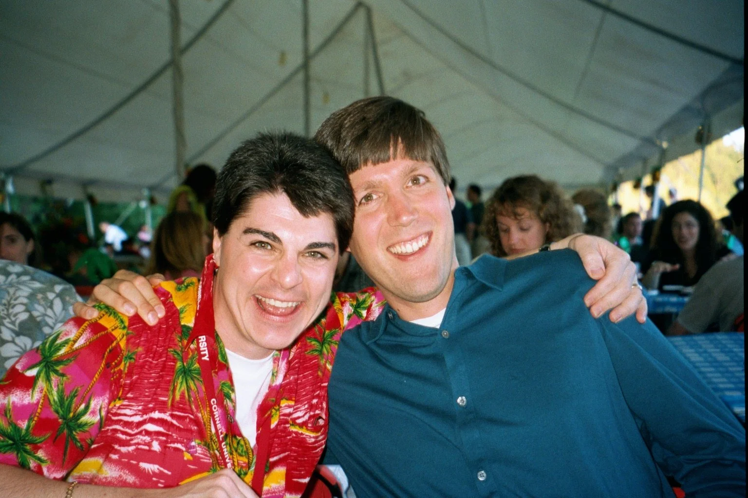 Two smiling men with their arms around each other at an outdoor event under a tent, with other people in the background.
