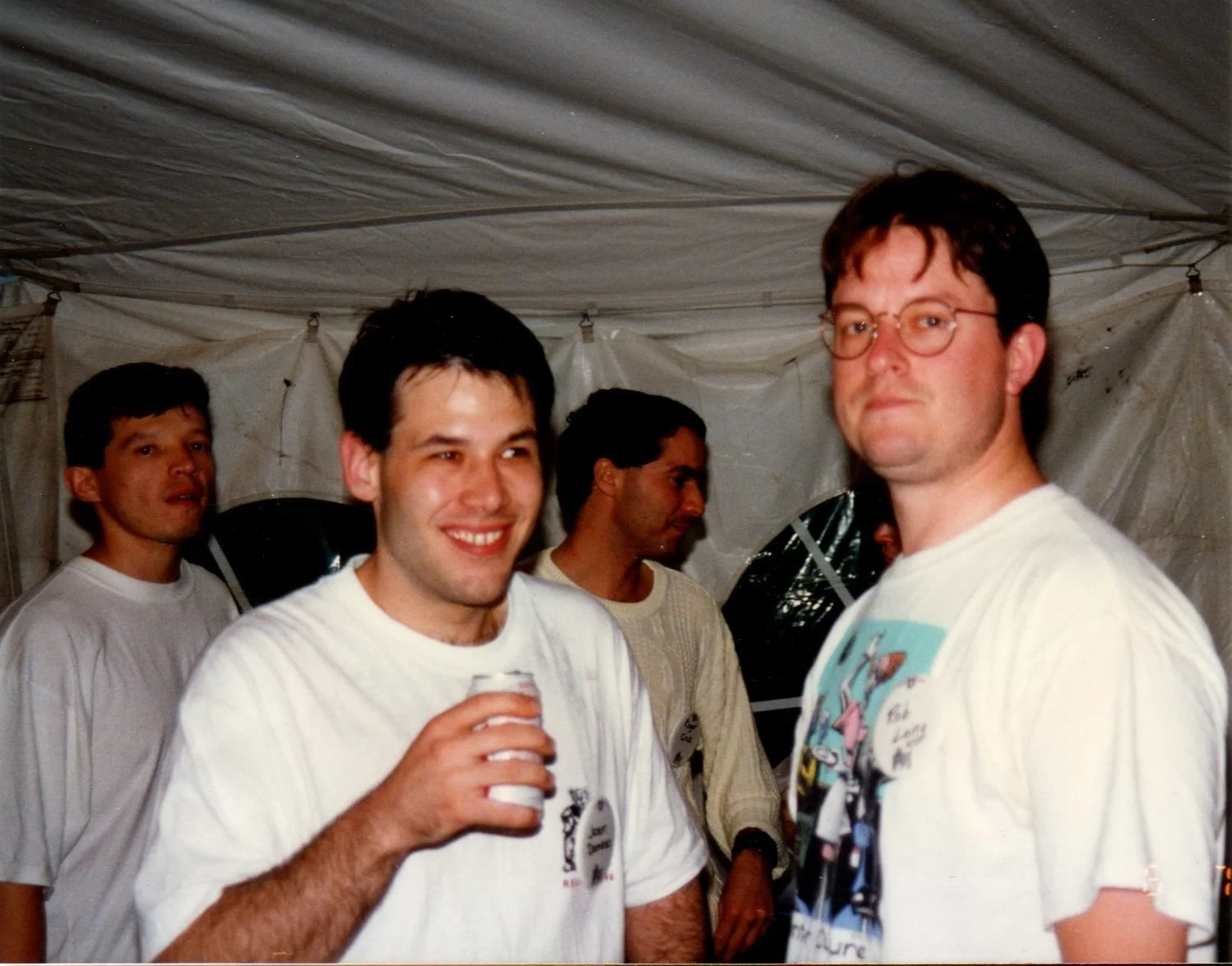 Group of five men standing inside a white tent at a social gathering, with two of them smiling at the camera.