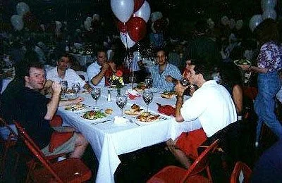 Group of young people sitting around a table at a celebration, with balloons and food, eating and drinking.