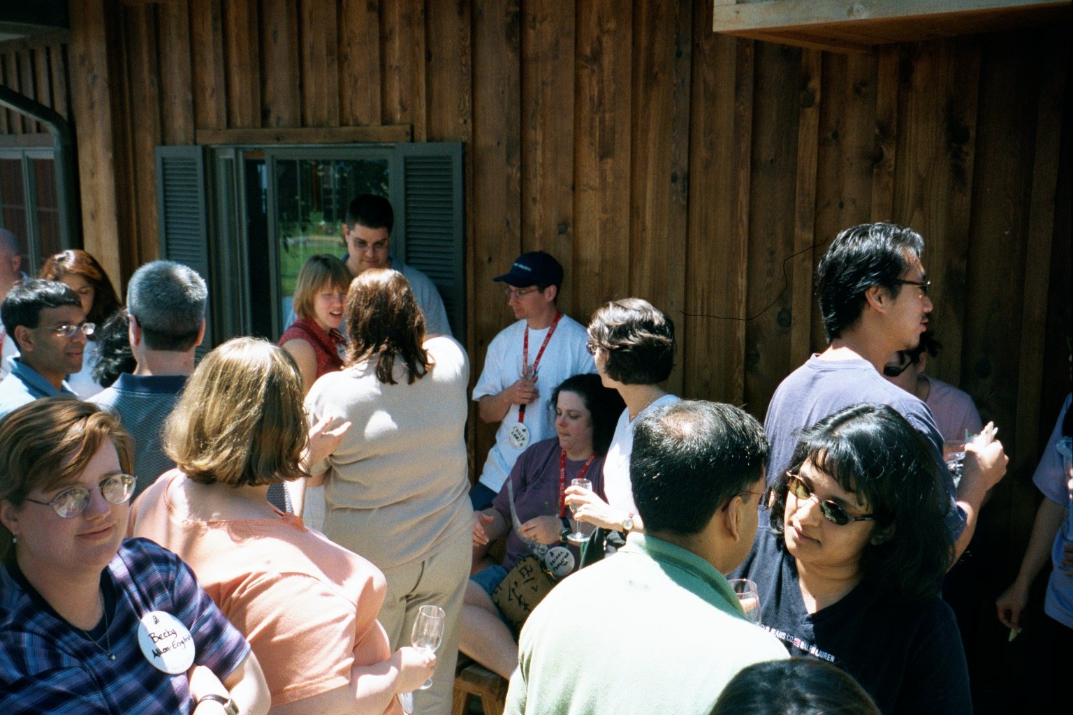 Group of people gathered outdoors in front of a wooden building, engaging in conversation and holding drinks.