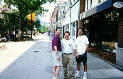 Three men standing on a city sidewalk, smiling for a photo, with trees and buildings in the background.