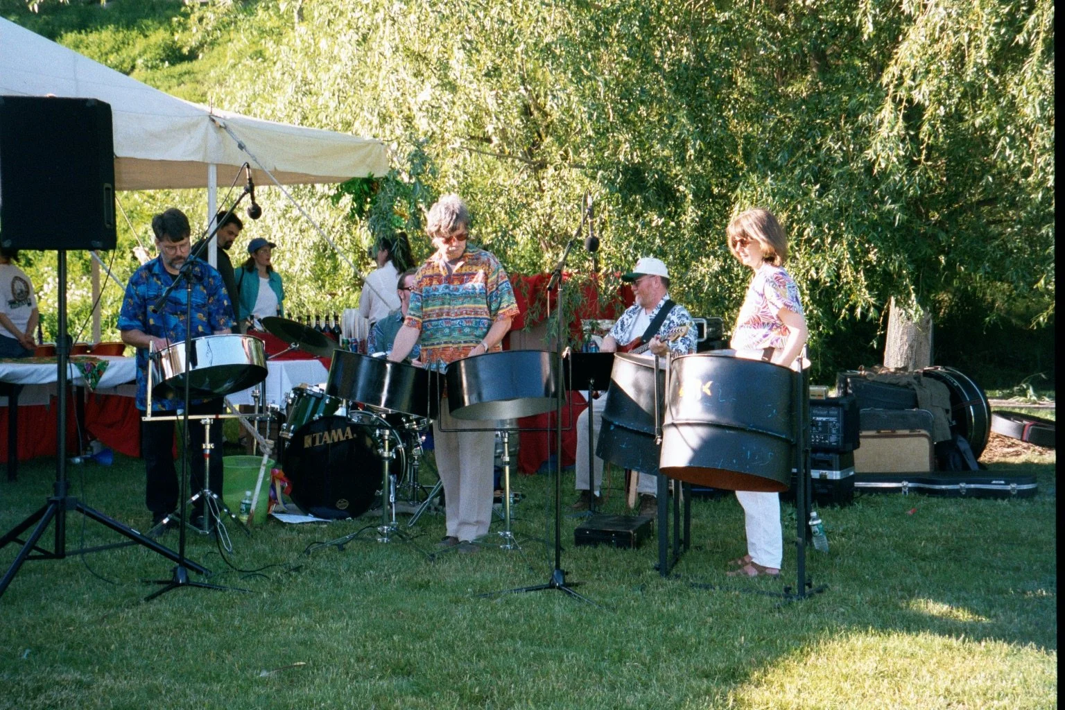 Band playing percussion instruments outdoors under a canopy at a sunny event, with people standing in the background and trees surrounding the area.