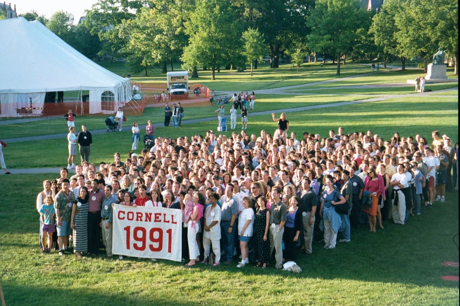 A large group of people gathered outdoors on a grassy area holding a banner that reads 'Cornell 1991.' There is a white tent, some trees, and a few people standing or walking around in the background.