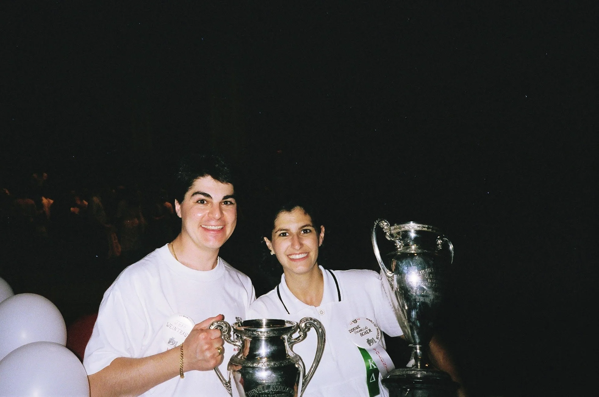Two women smiling and holding large silver trophies at a celebration event, surrounded by white balloons, at night under a dark sky.