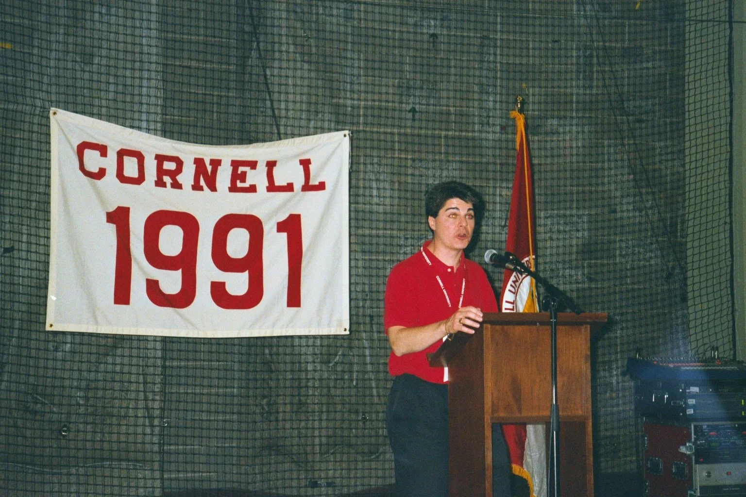 A woman speaking at a podium during a Cornell University event in 1991, with a Cornell banner and a flag behind her.