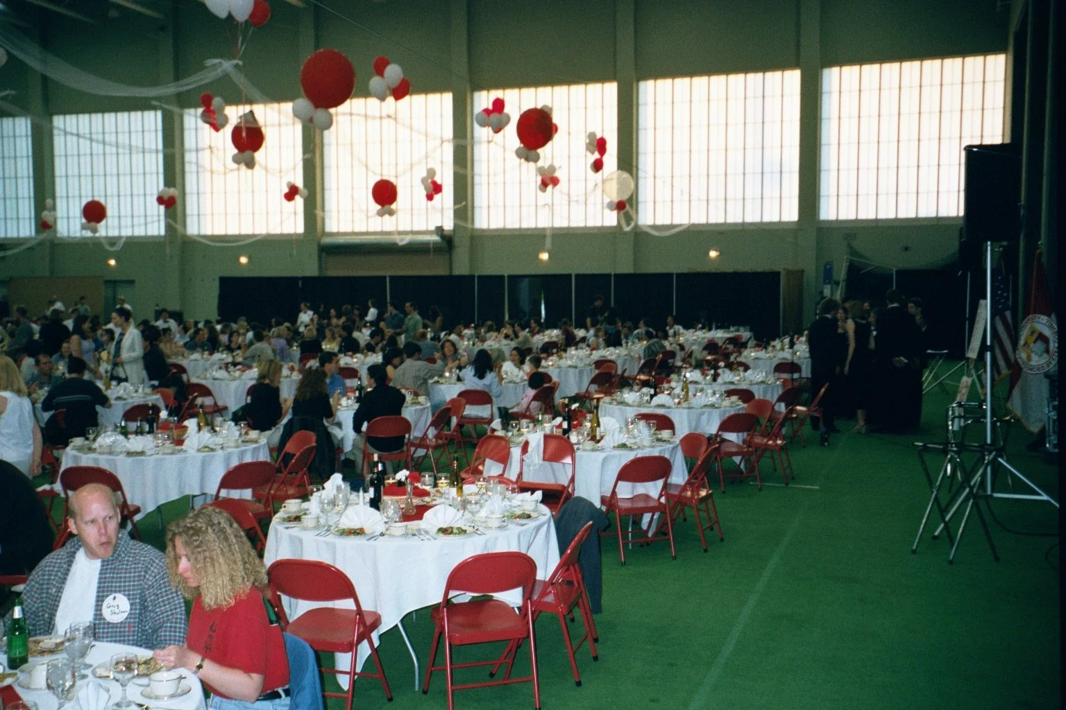 Indoor event with round tables covered in white tablecloths and red chairs, decorated with balloons and streamers, filled with people dining and socializing, some waitstaff serving, and a stage or speaker setup at the front.