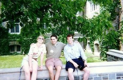 Three young adults sitting on a low brick wall outdoors, with green trees and a building in the background.