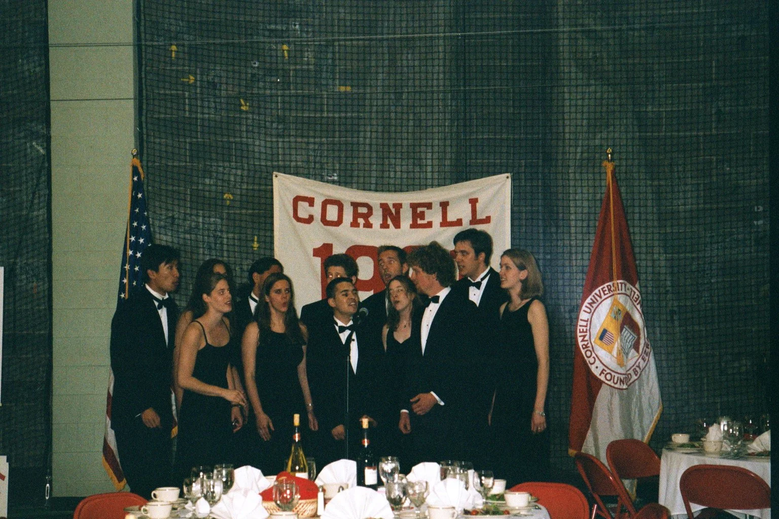 Group of young people in formal attire singing together at a Cornell University Reunion event, with American and Cornell flags in the background, and a large Cornell banner.