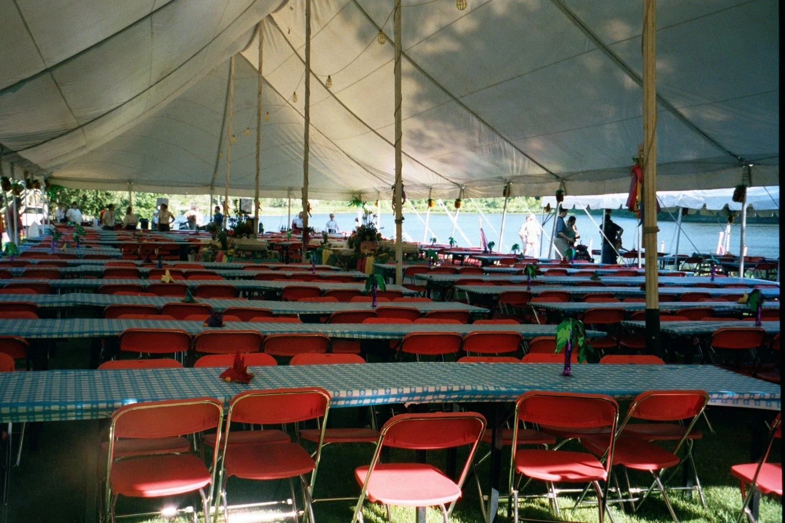 Large outdoor event under a white tent with several rows of red folding chairs and long tables covered with blue checkered tablecloths, decorated with small colorful ornaments, with people in the background near a body of water.