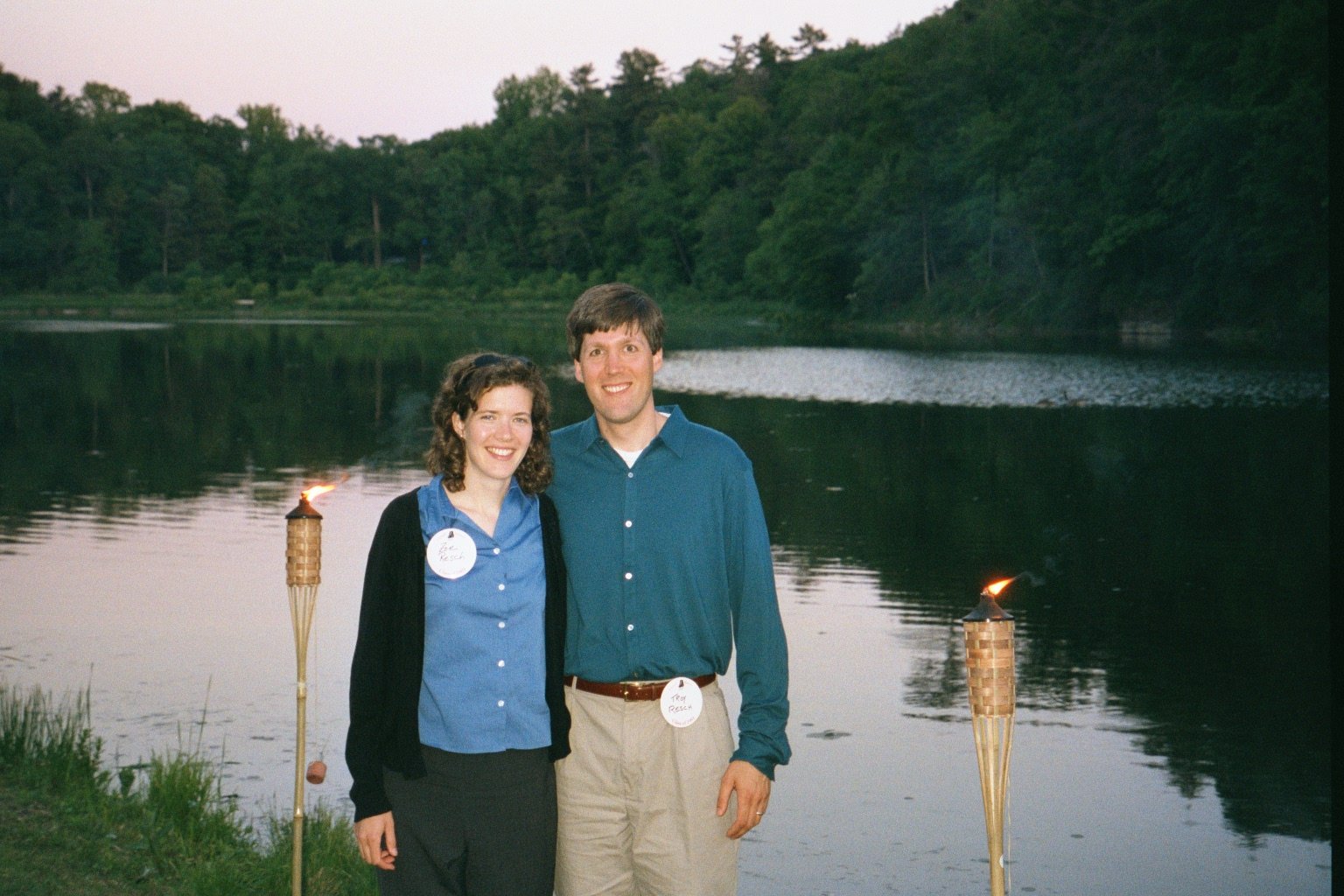 A smiling couple stands near Beebe Lake at sunset, with tiki torches, with a wooded area in the background during Cornell Reunion weekend.