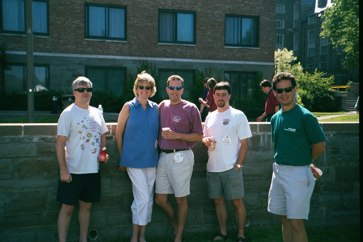 Group of five people standing outdoors, smiling, wearing casual summer clothing and sunglasses.