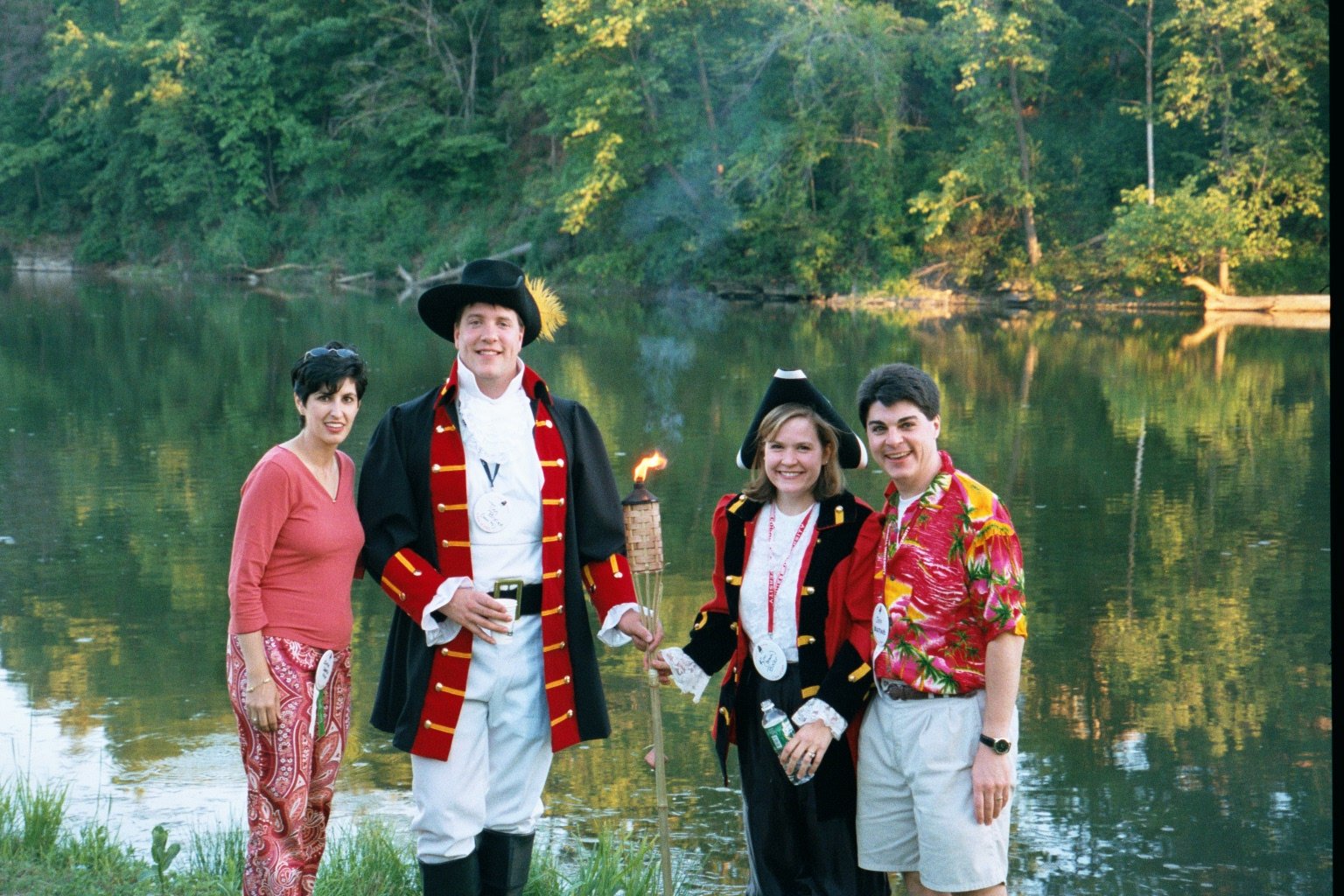 Group of four people dressed in historical and tropical clothing standing by Beebe Lake during Cornell Reunion weekend with trees in the background, one man holding a torch.