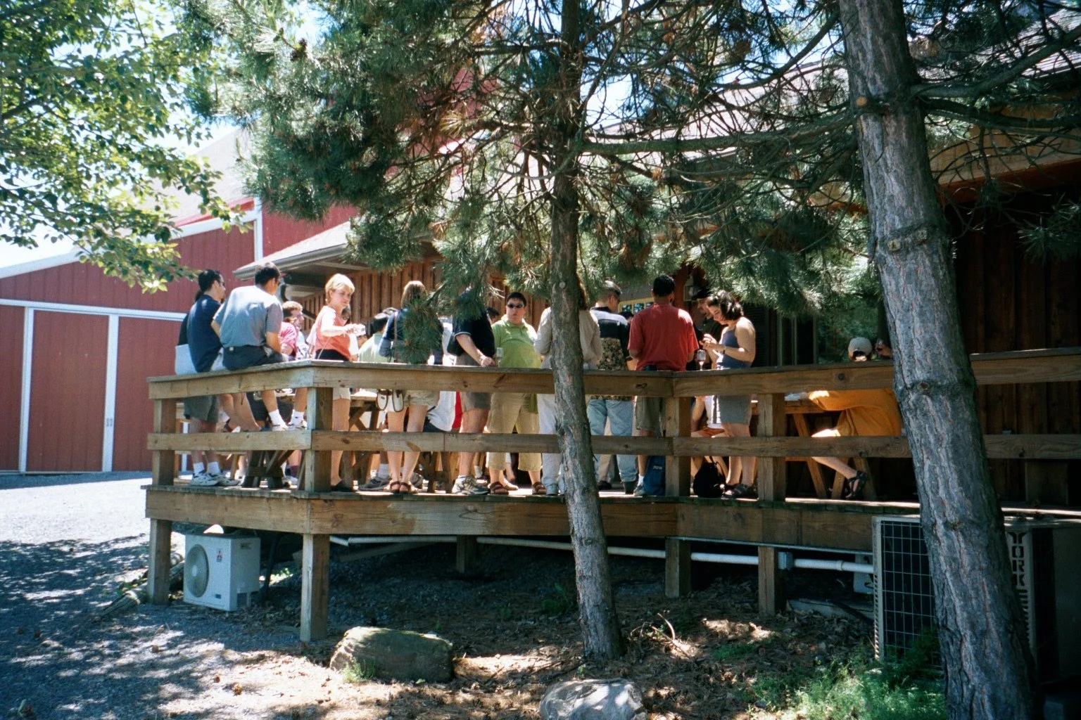 Group of people standing on a wooden deck beneath trees outside a red barn-style building on a sunny day.
