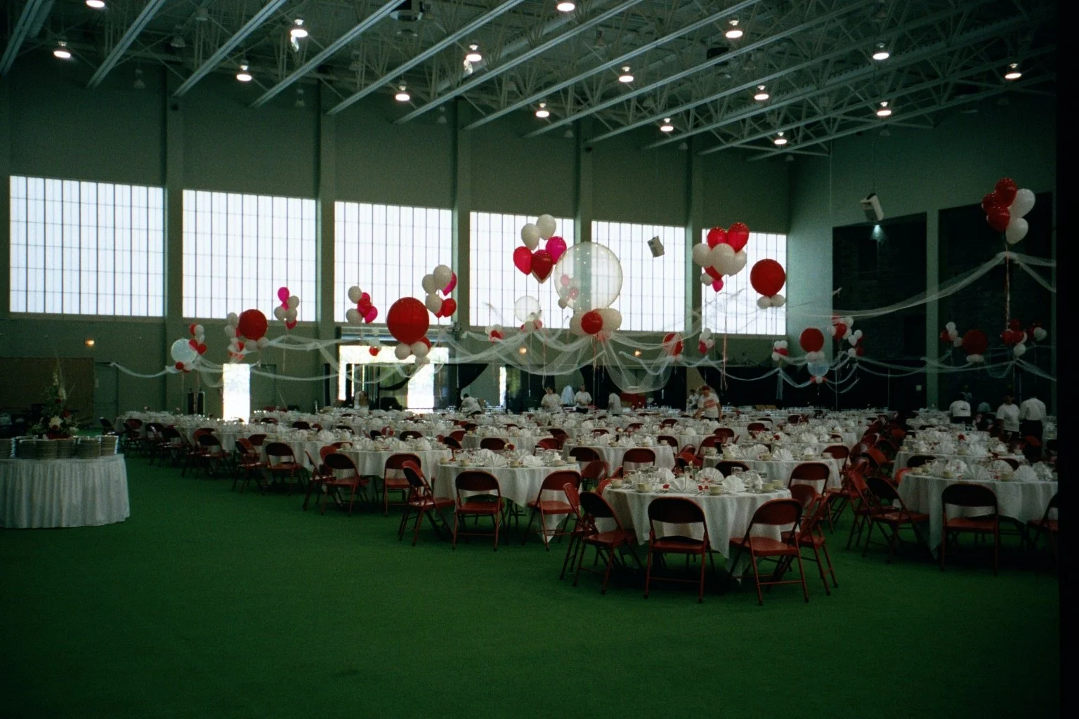 Indoor banquet hall decorated with red, white, and pink balloons, white tablecloths, and chairs arranged for a celebration.