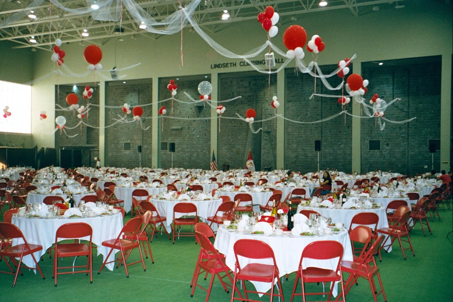 Banquet hall decorated for a celebration with red and white balloons and streamers, round tables with white tablecloths, and red chairs, set with plates, glasses, and bottles, with an American flag visible at the back.
