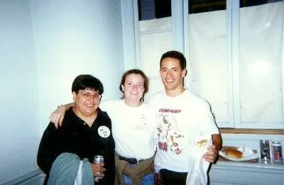 Three people standing together in a room with light-colored walls, smiling at the camera. The person on the left is wearing glasses and a dark hoodie, holding a can. The person in the middle is wearing a white shirt, and the person on the right is we