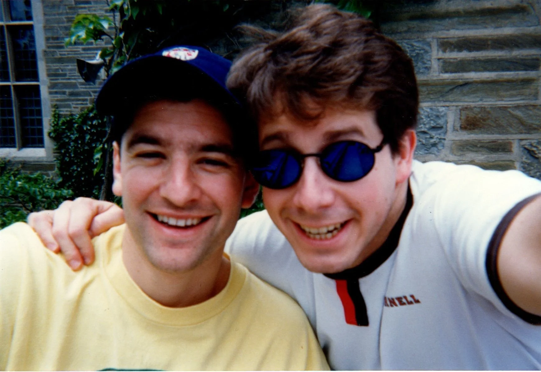 Two young men taking a close-up selfie outdoors, smiling and posing with arms around each other. One is wearing a yellow shirt and a dark cap, and the other is wearing reflective sunglasses, a white shirt with colored accents. There is greenery and a