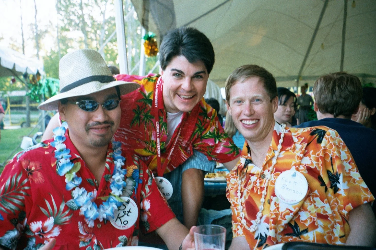 Three men wearing colorful Hawaiian shirts and necklaces during Cornell Reunion weekend, smiling at a festive gathering under a tent, with other people in the background.