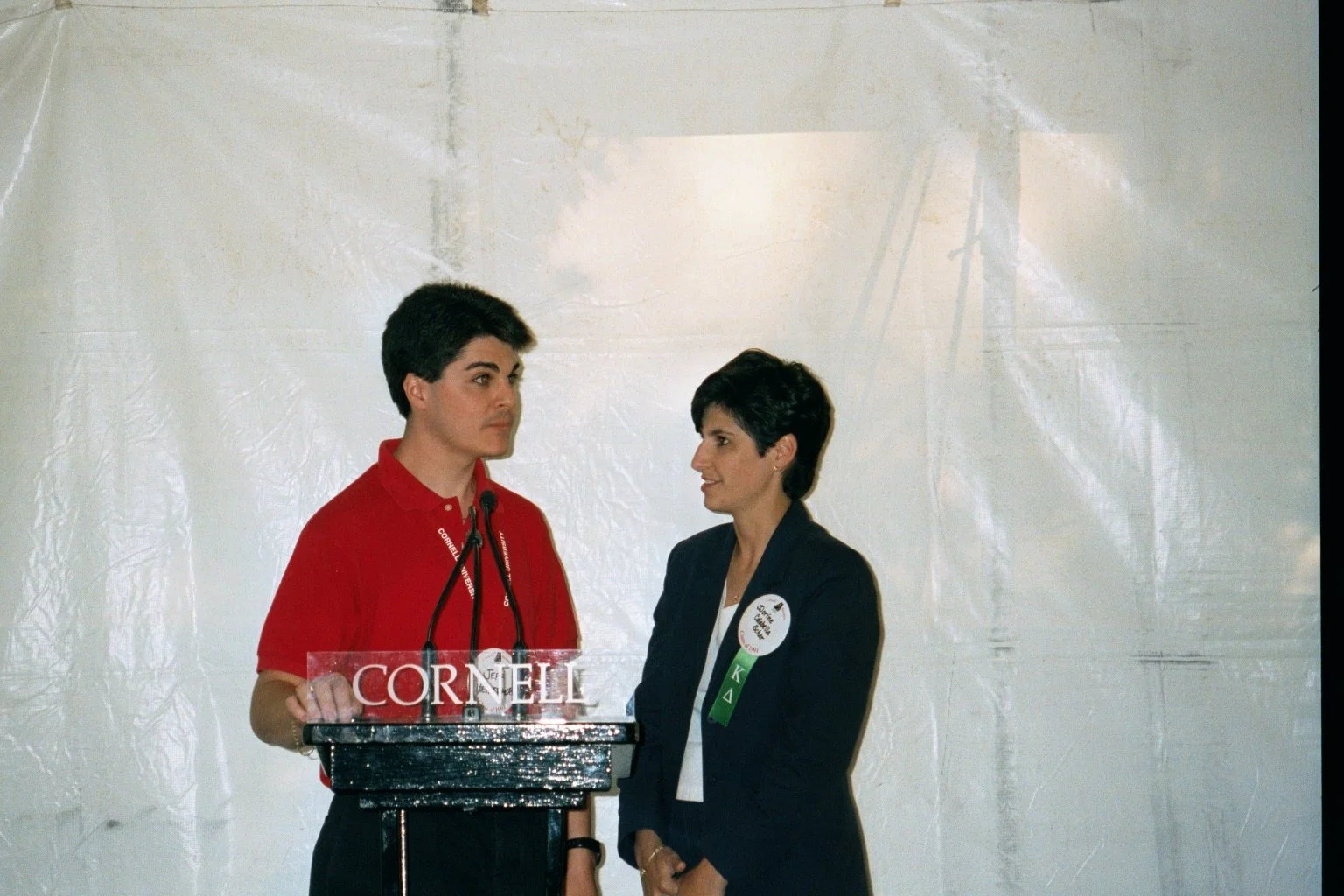 A man and woman standing at a podium with a Cornell sign, engaged in a conversation or speech, inside a large white tent.