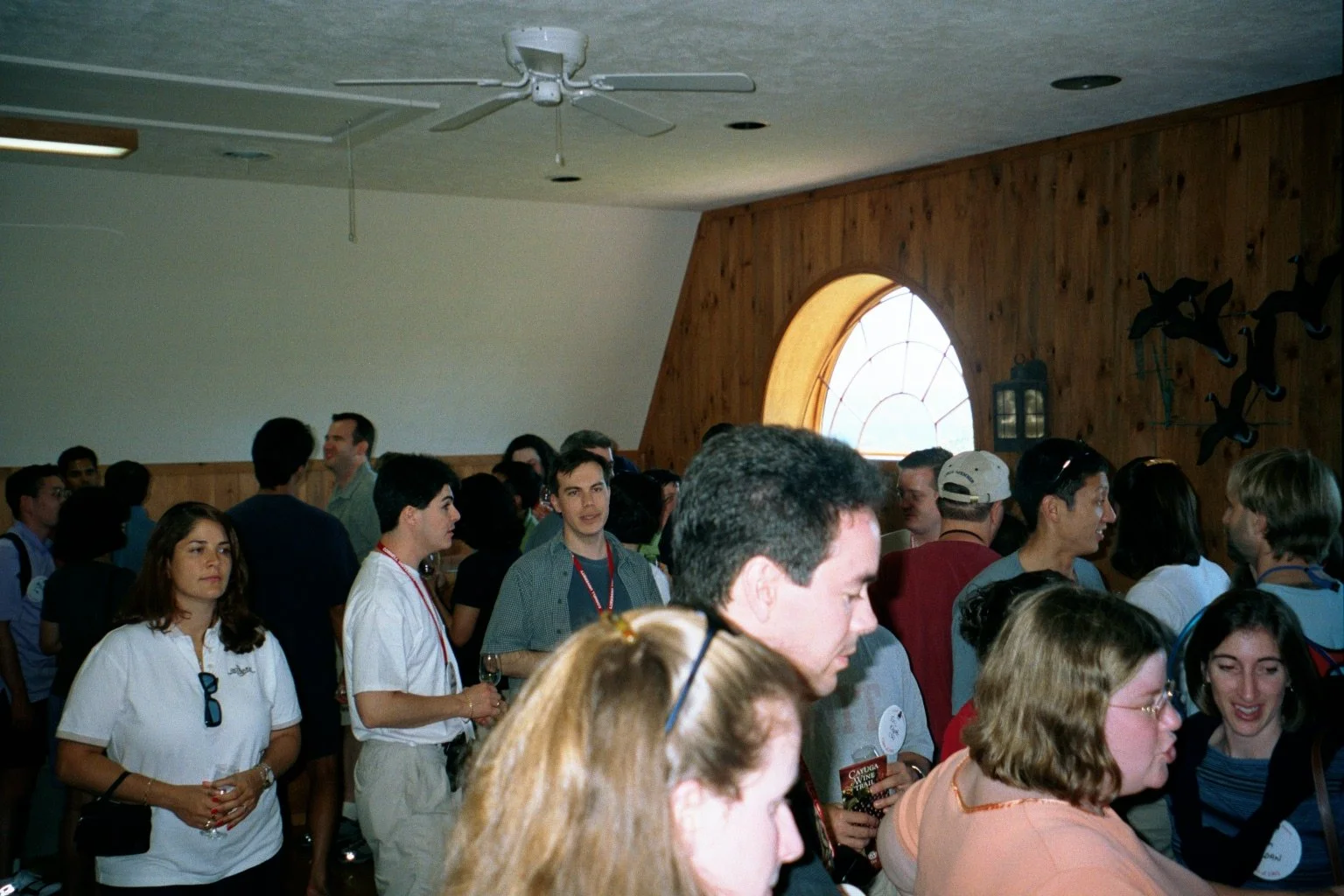 Indoor gathering of people, some talking and some standing in groups, with wooden walls and a round window in the background.