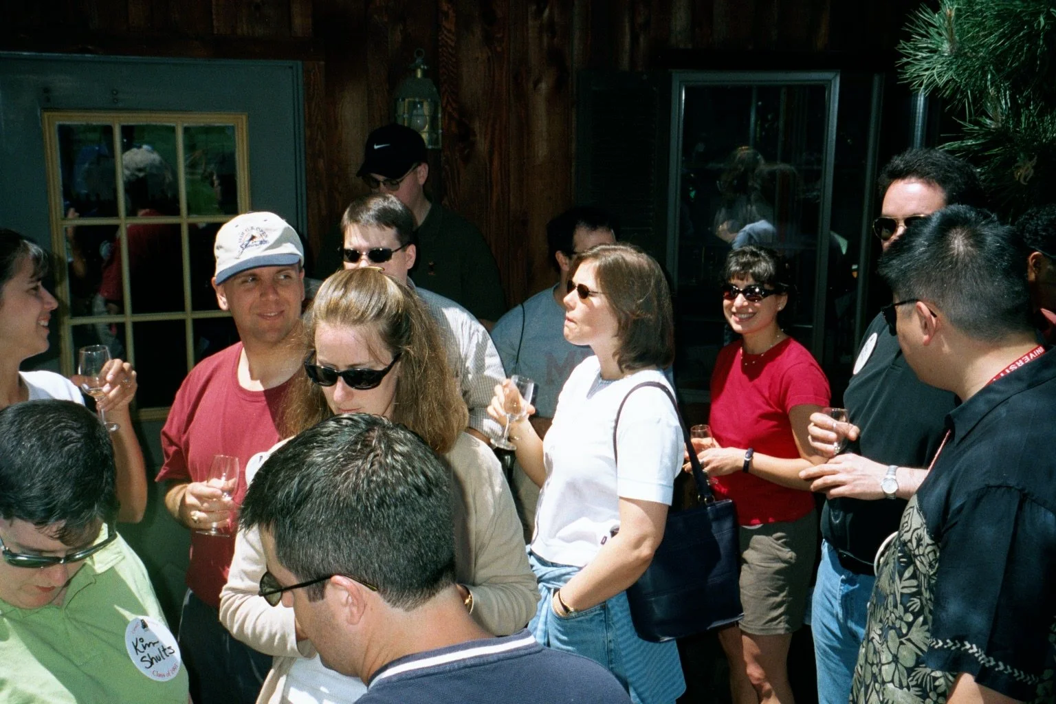 Group of people gathered indoors, holding wine glasses, engaging in conversation, with wooden walls and a window in the background.