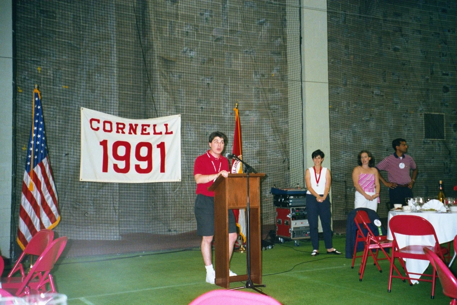 A woman speaking at a podium during a Cornell event in 1991, with an American flag and Cornell banner in the background, and people standing to her side in a large indoor space.