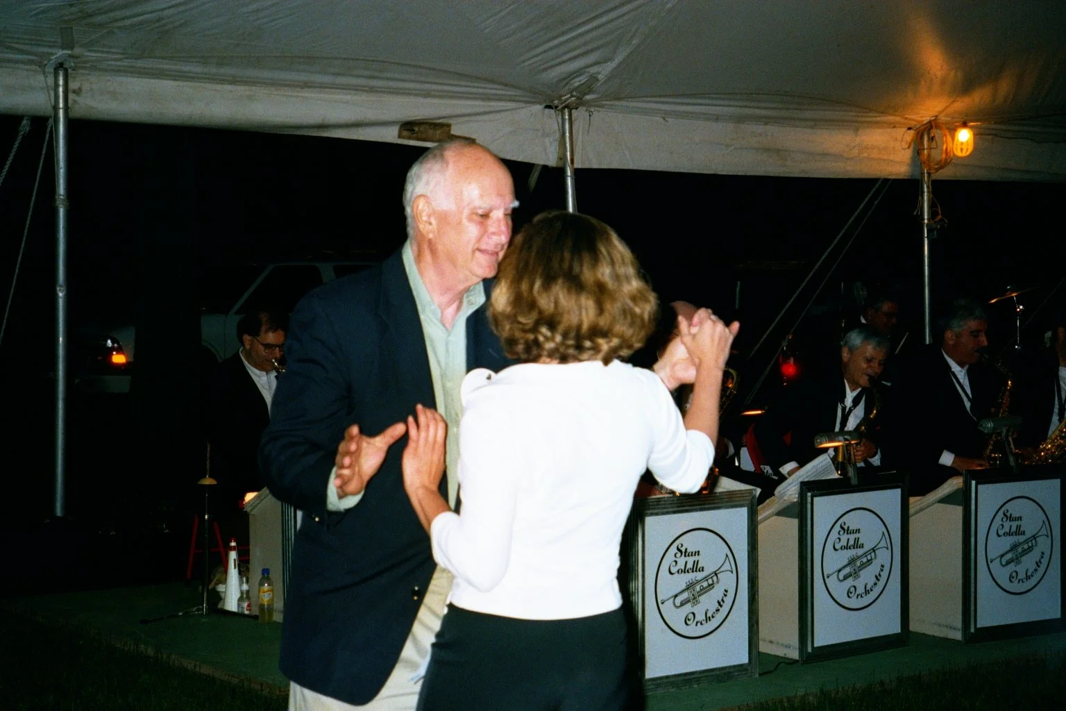 A man and a woman dancing in front of a band called Stan Colella Orchestra, playing instruments under a tent at night during Reunion weekend.