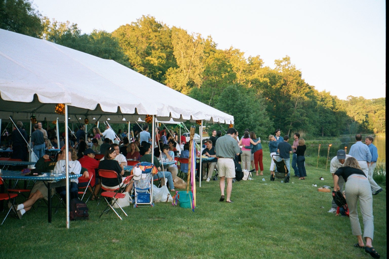 An outdoor gathering with a large white tent filled with people seated at tables, while others stand and socialize on the grass near a river, with green trees in the background during late afternoon or evening.