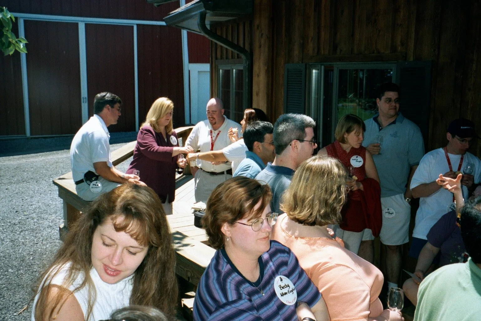 Group of people socializing outdoors, some are holding drinks and sitting on benches near a wooden building.
