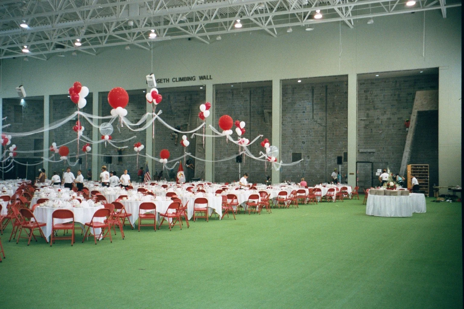 Indoor event space decorated with red and white balloons and streamers, with round tables and red chairs, and people preparing for an event.