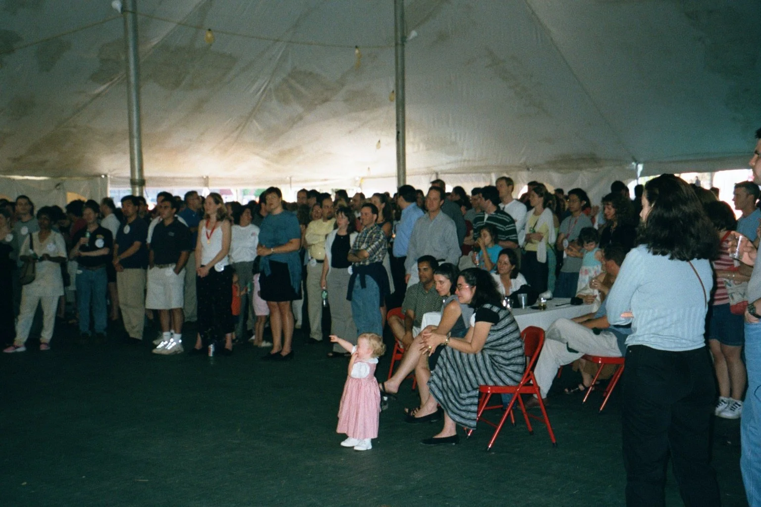 A large group of people gathered inside a tent, standing and sitting, watching an event with a young girl pointing towards something.