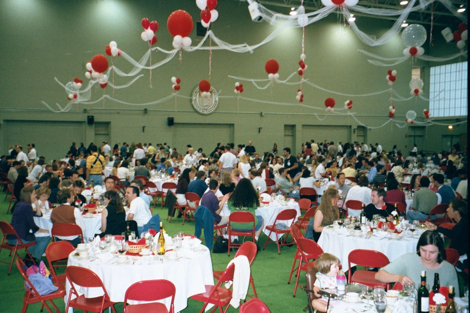 Large indoor banquet hall decorated with hanging white and red balloons and streamers, filled with numerous round tables occupied by people dining, with a large seal or emblem on the wall in the background.