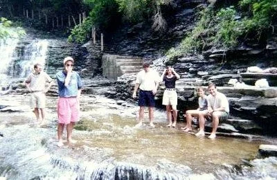Five people standing and sitting near a shallow stream in a forested area.