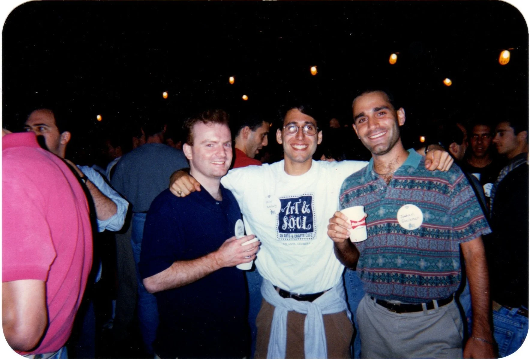 Three men smiling and posing for a photo at a crowded event, with most people in the background, at night with hanging lights.