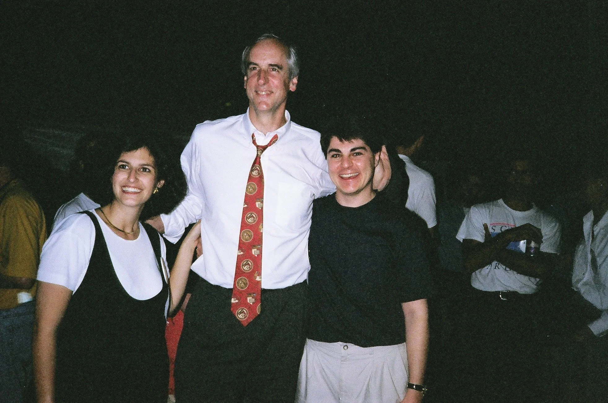 Three people standing together at night, smiling. The man in the middle is taller, wearing a white shirt and a red patterned tie, with his arms around a young woman on his left and a young man on his right. The young woman has dark curly hair and is