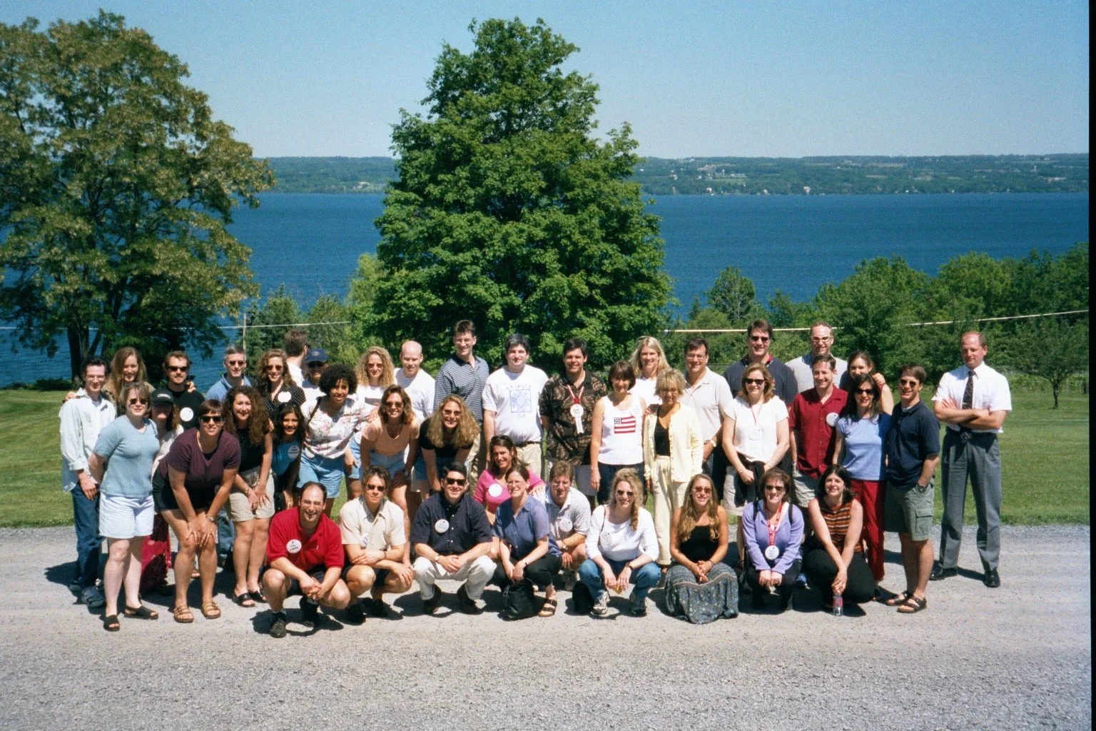 Group of people posed outdoors near a lake with green trees and blue water in the background, on a sunny day.