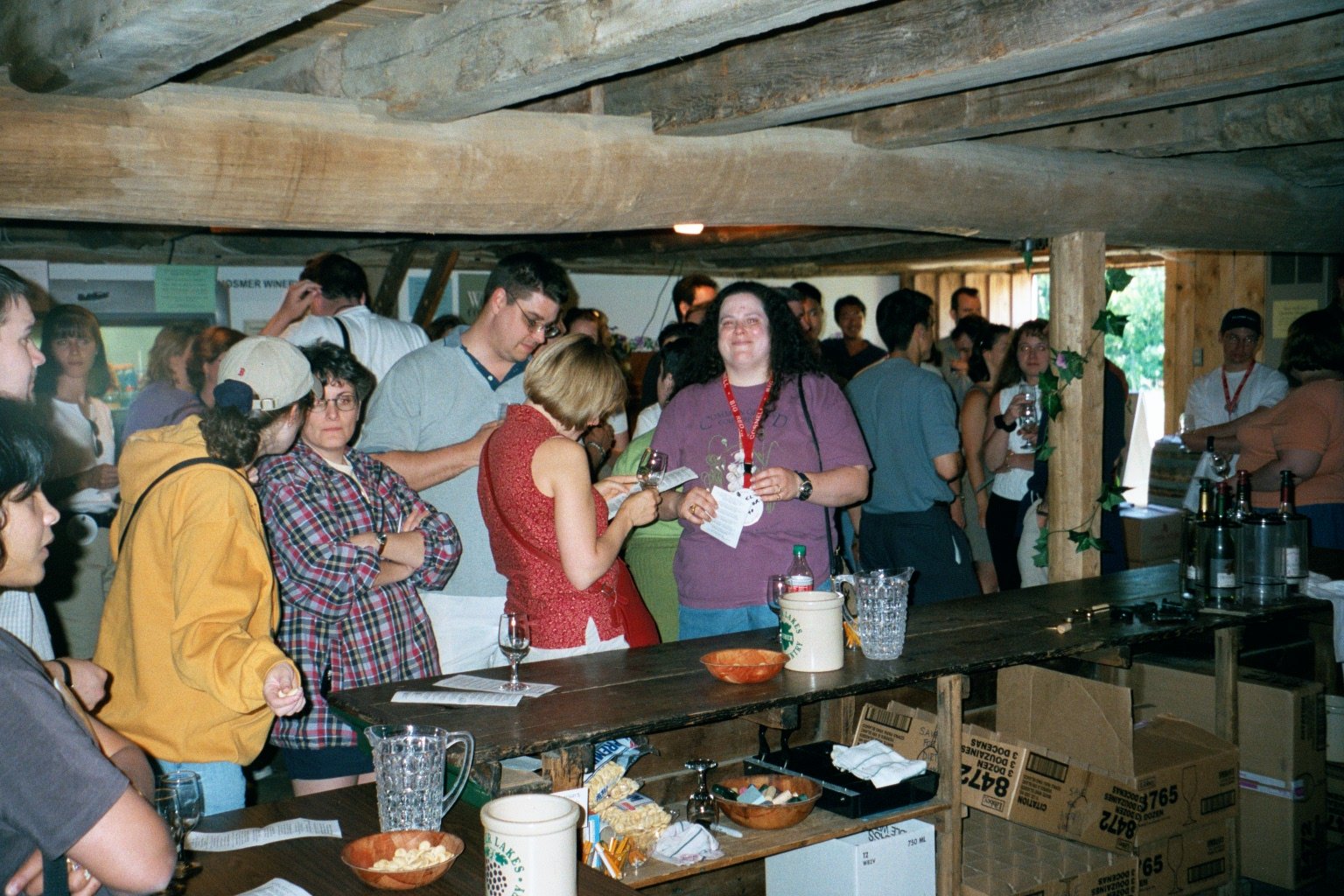 A group of people at a wine tasting event, standing in a rustic wooden room, with some holding glasses of wine, and a table with snacks and wine bottles in the foreground.