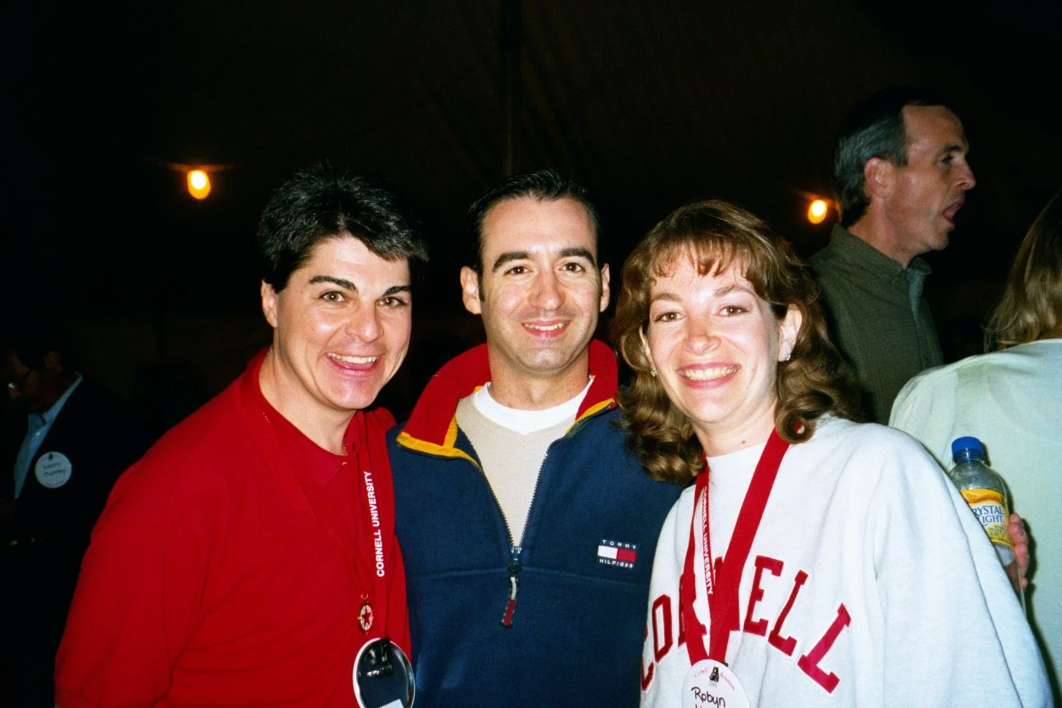 Three people smiling at a nighttime event, wearing casual clothing and red lanyards with name tags.
