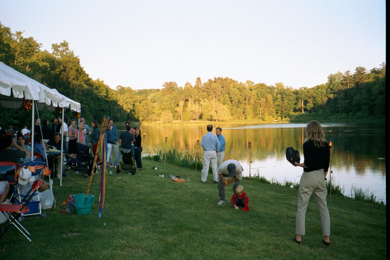 People gathered by a lakeside during a celebration, with some under a white tent, others standing and chatting near the water, and children playing on the grass, surrounded by trees and calm water at sunset.