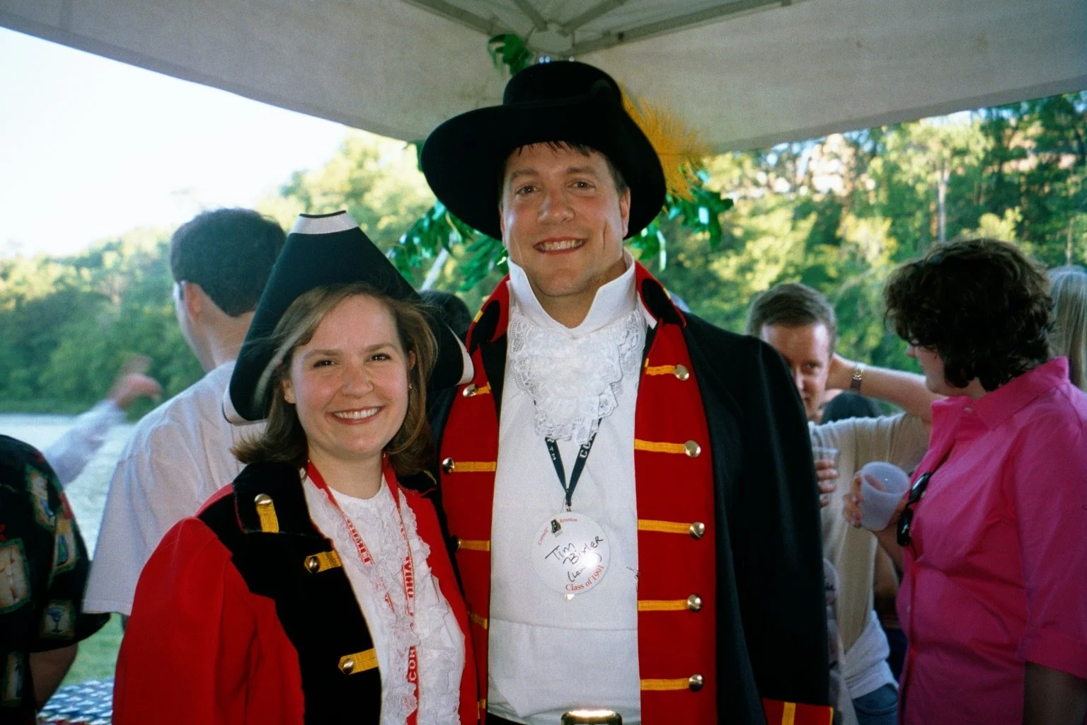 People dressed in costumes, with the man in the center wearing a historical military or pirate costume, and people at a social gathering outdoors.
