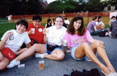 Four young friends sitting on a pavement outdoors, holding drinks and smiling, with other people sitting behind them.