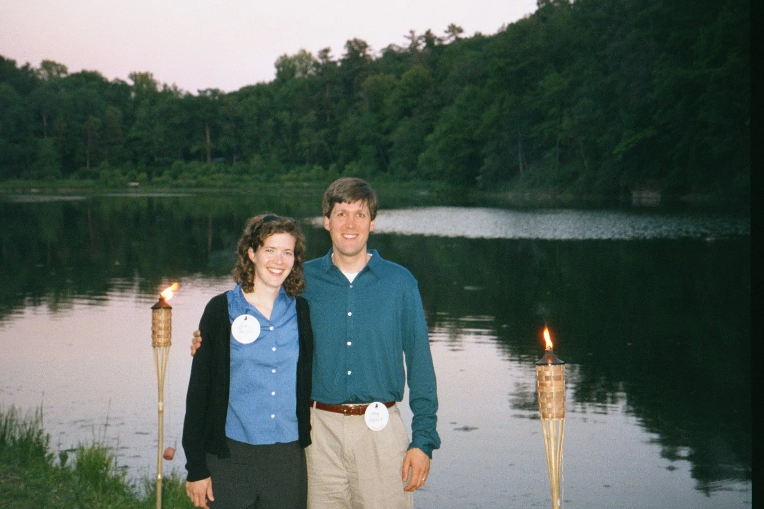 A smiling woman and man stand outdoors near a body of water, with torches on either side, and a lush green forest in the background during dusk.