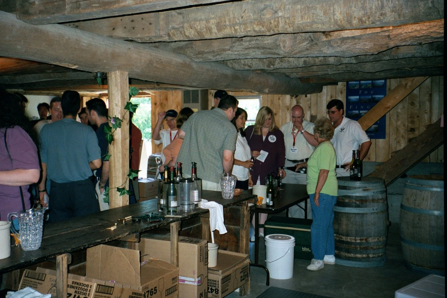People gathered at a wine tasting event inside a rustic wooden venue with barrels and boxes, engaging in conversation.