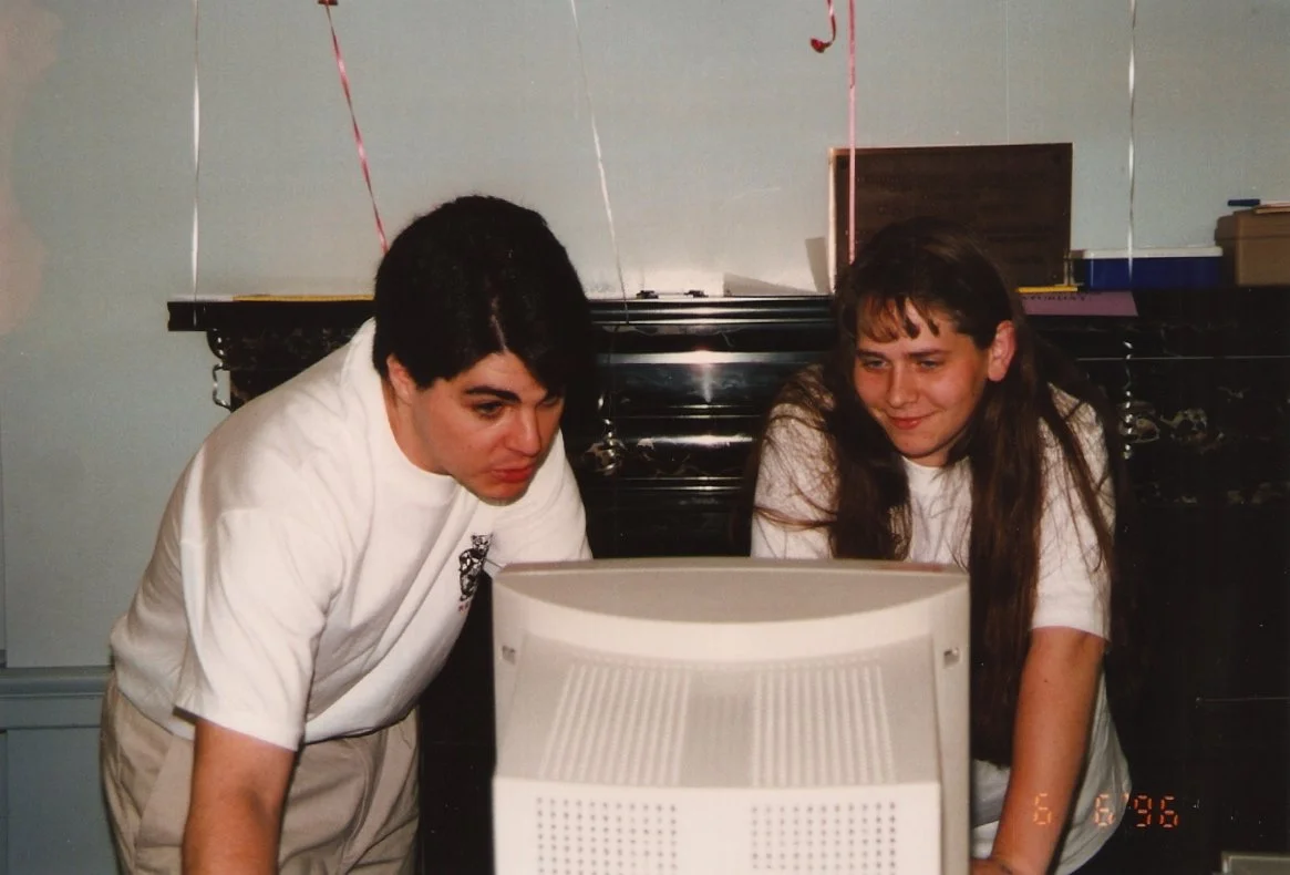 Two young women looking at an old desktop computer screen, with a black grand piano and some decorations in the background.