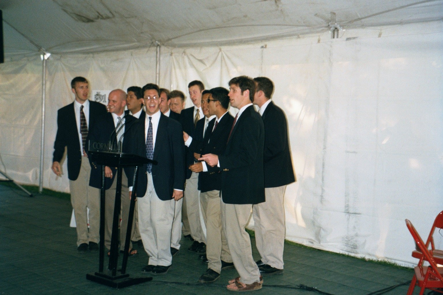 A group of men in business attire standing together at an event, some are smiling and talking, with a podium and microphones in front.