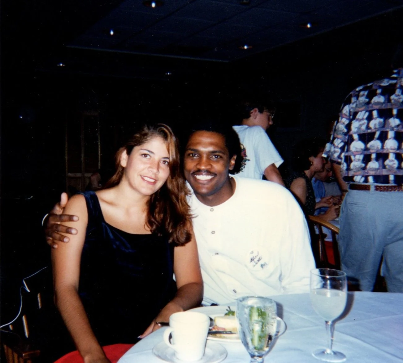 A woman and a man sitting at a dining table, smiling and posing for the photo. The woman has long, wavy brown hair, and the man is wearing a white shirt. There are teacups, glasses, and a plate on the table. In the background, there are other people