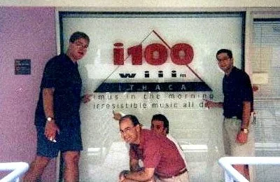 Four men in casual clothing standing in front of a wall with a radio station logo and slogan, some of them smiling or looking at the camera.