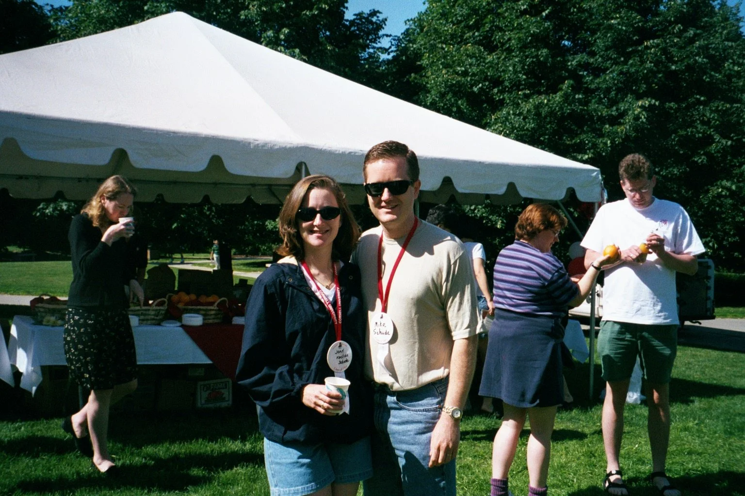 Two people standing outdoors in front of a white canopy tent, wearing sunglasses and name tags, at a gathering or party on a sunny day with trees in the background.